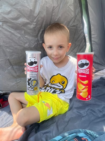 In Gagra, Abkhazia, a young boy sits inside a tent, holding two Pringles cans. The boy, with light brown hair and a white t-shirt featuring a yellow cartoon character, is positioned in the center of the frame. He is holding a silver Pringles can labeled "Mystery Flavour" and "Fallout 76" in his left hand, and a red Pringles can labeled "Original" in his right hand. He is wearing bright yellow shorts with cartoon shark graphics. The tent's interior is visible, with gray fabric forming the backdrop. The lighting suggests it is daytime.