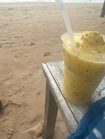 A clear plastic cup containing a yellow-green frozen beverage, flecked with dark particles and topped with a mound of the same mixture, rests on a weathered wooden surface, likely a chair or small table. A white, ridged plastic straw is inserted into the drink. The setting is a sandy beach in Tambon Ko Tao, Thailand, with the light brown sand exhibiting various footprints in the foreground and midground. A body of water, likely the ocean with faint waves, is visible in the background. A piece of blue fabric is partially draped over the wooden surface in the lower right quadrant. No individuals are visible.