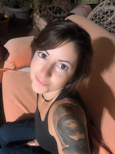 The image shows a close-up, overhead shot of a woman lying down outdoors. She has brown hair, dark eyes, and is wearing makeup. She is wearing a black tank top and has a large tattoo on her upper left arm. She is also wearing a dark beaded necklace and has a nose ring. The background appears to be a garden with potted plants and outdoor furniture, suggesting a residential setting within the Lachish Regional Council, Israel.