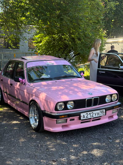 A pink, modified BMW sedan is parked outdoors on a gravel surface. The car has a sunroof and the sides are adorned with pink bows. A license plate reading "A 212 YH 98 RUS" is visible on the front. A banner with cartoon characters is affixed to the top of the windshield. In the background, a woman in a white dress is standing next to the open door of a dark-colored vehicle. A man is seated in the dark vehicle. Trees with green foliage are present in the background, and sunlight filters through the leaves. The environment appears to be a suburban or outdoor gathering space. The image does not depict Ladozhskoe Ozero, Russia.