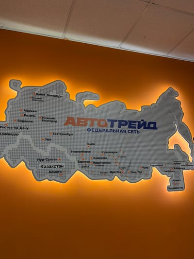 This media file showcases a decorative map of Russia with illuminated city names. The map is made up of a grey dotted pattern and is highlighted with a warm orange backlight, creating a visually striking effect against the darker orange wall. The prominent text "АВТОТРЕЙД ФЕДЕРАЛЬНАЯ СЕТЬ" is centered on the map, indicating it's likely a display for a logistics or transportation company.

Various Russian cities are marked with orange dots and labeled in Russian text, including St. Petersburg, Moscow, Yekaterinburg, Tomsk, Novosibirsk, Krasnoyarsk, Irkutsk, Ulan-Ude, Khabarovsk, and Vladivostok, among others. There are also labels for Kazakhstan cities like Nur-Sultan and Almaty.

The scene appears to be indoors, possibly in an office or public space, given the texture of the wall and ceiling visible on the left side of the image. The lighting suggests it could be daytime or a well-lit interior space, as there are no shadows indicating natural light sources. The overall mood is professional and modern, likely intended to represent the company's reach and network. There are no people visible in the image.