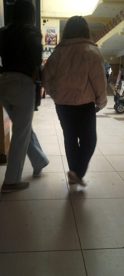 Two individuals are depicted walking through an indoor public space in Ngong ward, Kenya. The person on the left, viewed from the rear-left, wears a dark long-sleeved top, light-colored trousers, and light brown flat shoes. The person on the right, viewed from the rear, wears a light brown puffer jacket, dark trousers, and light-colored flat shoes. Both individuals are in motion, captured mid-stride on a light-tiled floor with dark grout lines. In the background, a white banner featuring "SPORTS GEAR" and graphic images is affixed to a light-colored wall. Further right, a multi-level interior structure is visible, characterized by a decorative railing on an upper floor and a small black wheeled vehicle in the distant background. The environment suggests a commercial building or shopping mall.