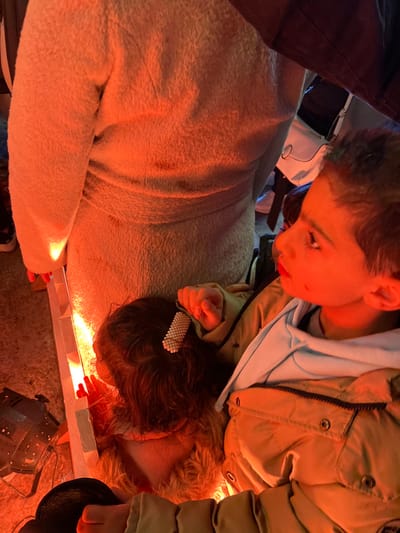 A young boy leans over, peering intently at something below him. He wears a khaki jacket over a light blue hooded sweatshirt. Beneath him, two people, possibly girls, are lying down, their faces close to a light source, possibly a makeshift stage or light box. One girl has a pearl-studded hair clip. The scene is lit with a warm, golden glow. A fuzzy blanket or coat drapes over one of the figures. The viewpoint is directly above, angled downward towards the subjects.