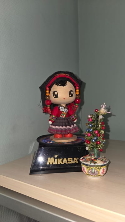 This media file features a decorative, handmade doll with a black headscarf and a red and black dress adorned with colorful embroidered patterns. The doll is wearing red shoes and has beads around its neck and ears. To its right, a miniature, beaded Christmas tree sits in a small, decorative bowl with floral patterns and Chinese characters. The doll and tree are displayed on a black base that appears to be a trophy or award, bearing the gold-colored text "MIKASA" and a star logo. The overall scene is brightly lit, with the subjects positioned against a plain, light blue background. The setting seems to be indoors, likely on a shelf or table, given the presence of a wooden surface to the right. The items suggest a festive or commemorative display. The visible text "MIKASA" indicates the possible origin or context of the display piece. The image does not provide clues about the time of day, weather, or specific emotions.