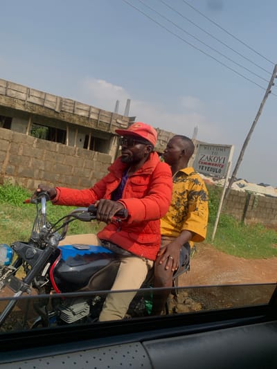 This media file captures two individuals on a motorcycle, with the shot appearing to be taken from within a vehicle. One person, wearing a red jacket and a red cap, is seated at the front, operating the motorcycle. They have glasses and a beard. Behind them, another person is seated, wearing a yellow shirt with a black pattern.
The scene is outdoors, with a dirt road and greenery visible in the background. Construction is also evident, with a building under construction featuring brickwork and visible rebar. Overhead, power lines are present against a bright sky, suggesting daytime. The weather appears to be clear and sunny.
A sign is visible that reads "WELCOME TO ZAKOYI COMMUNITY YEYEDULO". The overall impression is one of an everyday scene in a developing area, possibly in Dutse, Nigeria, given the provided location context. The subjects do not display strong emotional expressions, appearing focused on their journey.