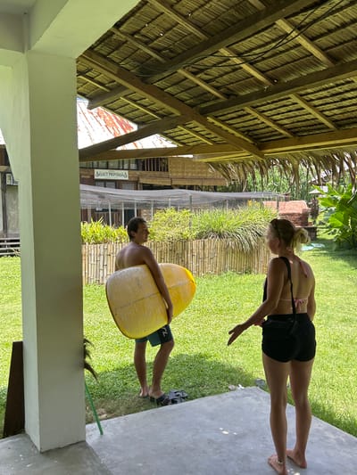 A woman and a man stand in a grassy yard near a building. The man, carrying a yellow surfboard, is positioned closer to the building. The woman stands further away and appears to be looking towards the man. The building is a structure with a wooden frame and a thatched roof. The location is undetermined as the city of Santa Monica is not in the Philippines.