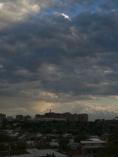 The image depicts an urban landscape under a dramatic sky. Heavy, dark storm clouds dominate the left side of the frame, contrasting with a lighter, brighter area on the right, suggesting the setting or rising of the sun.

Below the sky, there is a view of a town. Buildings of various sizes are visible, interspersed with trees and greenery. There appears to be a construction crane in the scene, indicating ongoing development.
In the distance, there are hints of mountains. The city of Arinj, Armenia has views of Mt. Ararat, so it is likely that those are the mountains in the background.