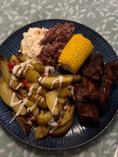 The image presents a close-up, overhead view of a single plate of food, positioned on a light teal-colored, patterned textile surface within an indoor setting. No individuals are visible.
The circular dark-blue plate features a textured, radially ribbed rim. The plate holds a composed meal consisting of five distinct components:
1. **Roasted Potato Wedges**: A pile of irregularly cut, golden-brown potato pieces, some with skin intact, intermixed with small red diced vegetables (likely bell peppers) and green herbaceous flakes. These are uniformly drizzled with a thick, white, opaque sauce applied in zigzag lines.
2. **Barbecued Ribs**: Approximately four dark-brown, glossy meat segments, consistent with pork ribs, coated in a dark, viscous barbecue sauce, exhibiting some areas of charring.
3. **Corn on the Cob**: A single half-segment of cooked yellow corn on the cob, displaying visible, distinct kernels.
4. **Coleslaw**: A serving of creamy white and orange shredded vegetables, consistent with coleslaw.
5. **Shredded Preparation**: A mass of finely shredded, reddish-brown material, consistent with pulled meat or a red cabbage slaw, appearing moist.
The underlying surface is a textile, possibly a blanket or tablecloth, in a light teal or mint green hue, adorned with a repeating pattern of lighter-toned, abstract floral or leaf motifs. The specific location within New Forest, United Kingdom, cannot be determined from the visual information provided in the image.