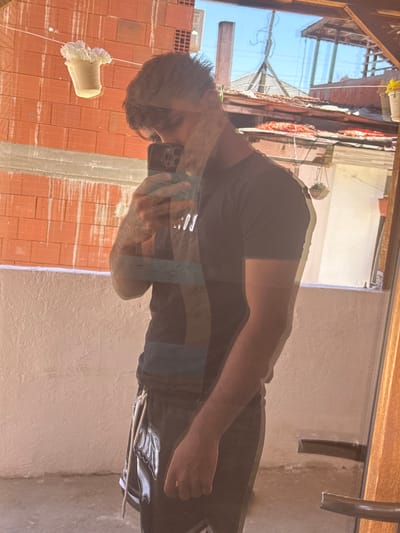 A mirrored image of a man taking a photo with his phone is reflected in a window. The man has short dark hair and is wearing a dark T-shirt and dark patterned shorts. He is standing in front of a brick building under construction, partially covered with plaster. Outside the window, the sky is blue, and a glimpse of houses with terracotta roofs is visible. The window frame is brown. The mirrored reflections suggest a residential area or construction site in Sandanski, Bulgaria.