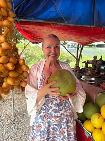 A Caucasian woman with blonde hair pulled back smiles at the camera, holding a large, green pomelo in her arms. She is wearing a white robe with intricate gold-toned patterns over a floral-print dress. The woman stands at an outdoor market stall in Rai Mai, Thailand, as suggested by the abundant tropical fruits visible. To her left, a cluster of ripe, golden dates hangs from a stalk. Behind her, a shelf displays various small statues and trinkets. The stall is covered by a blue and red awning. The ground is covered with small, grey pebbles. In the background, lush green foliage and trees are visible, with a hint of a fence and structures beyond.
