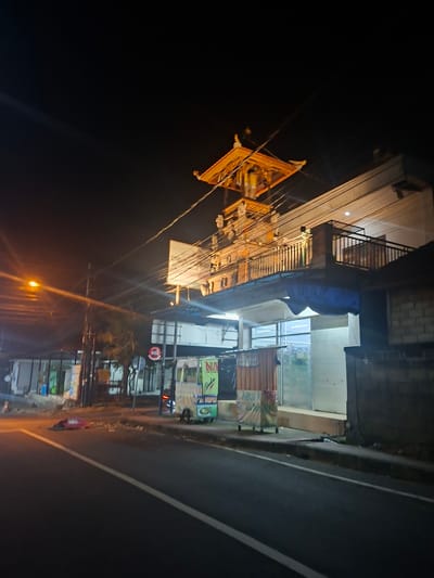 The image shows a street scene at night in Blahbatuh, Indonesia. A street light illuminates the asphalt road and the side of a building. The building appears to be a shop or a small business, with goods displayed in windows and on shelves. There are signs with text in what appears to be Indonesian. In the foreground, a yellow and orange structure, possibly an altar or a decorative piece, hangs from wires. The overall atmosphere is that of a busy street at night, with lights and activity. The image is slightly tilted, giving a dynamic perspective.