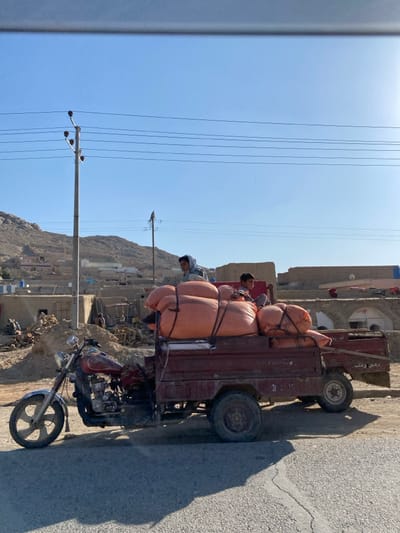 This image depicts a three-wheeled motorized cargo vehicle, commonly known as a motor-rickshaw or cargo tricycle, parked or in motion on an asphalt road in کوکران, Afghanistan. The vehicle consists of a reddish-brown motorcycle front section connected to a rear cargo bed of the same color. The cargo bed is heavily loaded with approximately six large, orange, bulging sacks, secured with black straps. Two individuals are seated on top of these sacks: an older male, likely a teenager or young adult, with a light-colored head covering, positioned towards the front-left of the cargo; and a younger boy in a dark shirt, seated behind him towards the right.
The scene is set under a clear, bright blue sky, indicating daytime. The background reveals a semi-arid, rural or semi-urban landscape characterized by low-rise, earthen-toned structures, possibly mud-brick or adobe, some appearing to be in various states of repair or construction. To the left, a significant pile of earth or construction debris is visible near several individuals in the midground. Rocky, barren mountains rise in the far background. Two utility poles with multiple overhead wires are prominent, one on the left in the midground and another further back on the right. The road in the foreground exhibits a visible crack, and the vehicle casts a distinct shadow to the right.