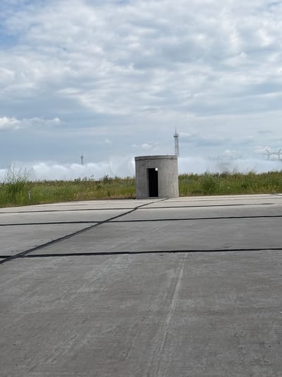 The image depicts a view of the territory surrounding the South Ukrainian Nuclear Power Plant in the city of Pivdennoukrainsk, Ukraine. In the foreground, a concrete surface with visible seams is visible, suggesting a paved area like a road or a helipad. A circular concrete structure with an open doorway stands in the middle ground. Behind this structure, a line of green vegetation borders a field. In the background, the sky is filled with white, fluffy clouds. A tall, slender tower with an antenna is visible on the left side of the background. To the right of the tower, a large plume of white mist or spray is emanating from the ground, obscuring some of the background infrastructure. Power lines and transmission towers are partially visible through the mist in the far distance. The overall scene appears to be an industrial or utility site on an overcast day.