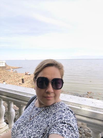 A woman wearing sunglasses and a floral top takes a selfie outdoors. She is standing on a paved walkway behind a white railing, with a view of the Gulf of Finland. The background shows a rocky shoreline with several people sitting and standing. In the distance, a tall building, likely the Lakhta Center, is visible on the horizon. The sky is overcast. The image appears to be taken in Peterhof, Russia, a suburb of Saint Petersburg, known for its imperial palaces and gardens.