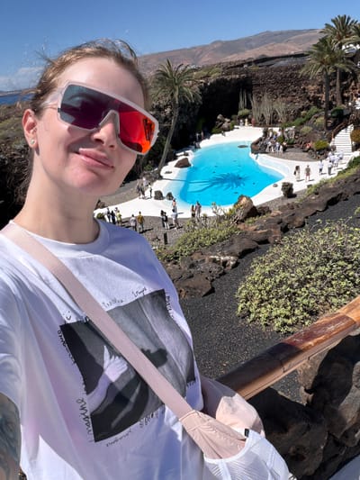 A selfie captures a person wearing sunglasses, a white t-shirt with a graphic print, and a light-colored shoulder strap or bag. The person is positioned to the right in the frame. The background is a complex, scenic landscape, likely in the city of Haría, Spain. There's a bright blue, irregularly-shaped pool with palm tree reflections on its surface. Around the pool, there is a white border and a dark, volcanic rock landscape. In the distance, there is a light brown mountain. People are visible around the pool area, suggesting it is a public or tourist attraction. A wooden railing is on the right side of the frame, slightly out of focus.