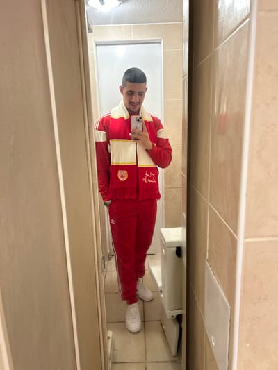 A man stands in a tiled bathroom, facing a mirror. He wears a red tracksuit with white stripes and white sneakers. A red and white scarf with fringe is draped around his neck. He is holding a phone up, presumably taking a selfie. A toilet and other bathroom fixtures are visible in the reflection. The room is lit by fluorescent lighting.