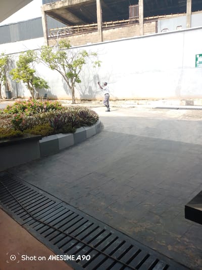 A man in a white shirt and grey pants, wearing a cap, is captured in mid-air as he jumps or leaps. He is standing on an asphalt surface, with a white wall behind him, and trees casting shadows on the wall. In the foreground, there is a paved area with a metal grate and a planter with lush green plants and pink flowers. To the left of the planter is a dark, bench-like structure. In the background, there is a multi-story building under construction, with exposed concrete and scaffolding. The scene suggests an outdoor setting, potentially a public space or a commercial area in Ibadan, Nigeria, given the specific details of the building and surrounding environment. The lighting indicates it is daytime.