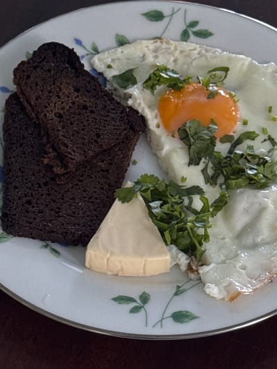 A fried egg with a runny yolk is served on a white plate with a decorative border. The egg is garnished with chopped herbs, possibly cilantro. To the left of the egg are two slices of dark rye bread, stacked on top of each other. A wedge of pale yellow cheese, possibly cream cheese or butter, is placed between the bread and the egg, also garnished with herbs. The plate is adorned with a pattern of green leaves and blue flowers. The background suggests a dark wooden surface, possibly a table. The image is an overhead shot of the food on the plate. The location is not discernible from the image.