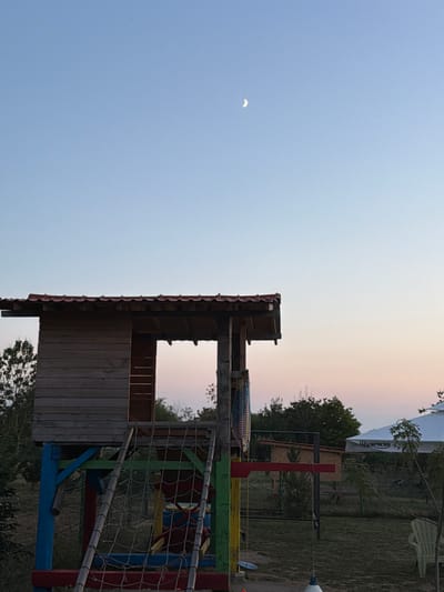 The image captures an outdoor scene at dusk in Tvarditsa, Bulgaria. The foreground features a brightly colored wooden playground structure, characterized by its red tiled roof and blue, red, green, and yellow painted supports. A rope net ladder extends from the structure. To the right, a white umbrella with Cyrillic text is visible, partially obscuring a smaller wooden building behind it. In the distance, trees are scattered across the landscape, and the sky is a gradient of pale blue and soft pink, with a crescent moon visible in the upper portion of the frame. The ground is covered in grass. The overall atmosphere is serene and twilight.