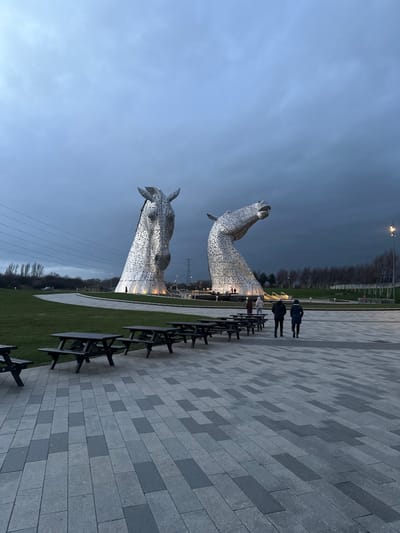 The image displays an outdoor scene at The Kelpies, located in The Helix park, Falkirk, United Kingdom, under an overcast, dark grey sky. Two monumental, metallic horse-head sculptures, illuminated from below, dominate the mid-ground. The sculpture on the left faces forward, while the one on the right has its head raised and turned. In the foreground, a paved area with grey and darker grey rectangular pavers features a linear arrangement of at least seven dark-colored wooden picnic tables with attached benches. Four individuals are walking away from the viewer towards the sculptures in the mid-ground; two in dark jackets are in the foreground of this group, with two others further back. Multiple smaller figures are visible near the base of the sculptures. A curving expanse of green grass extends from the paved area towards the sculptures. Overhead power lines are visible on the left side, and bare-branched trees line the background on the right. A single street light stands on the far right.