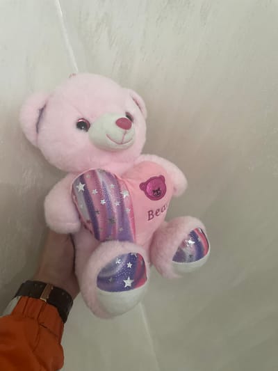 A pink plush teddy bear is depicted, held by a human hand against a light, plain background, likely an interior wall. The teddy bear features shiny pink glitter in its eye area and a pink heart-shaped nose. It holds a heart-shaped pillow in its arms; the left side of the pillow displays vertical stripes in shades of pink, purple, and blue, adorned with white stars and glitter, while the right side is solid pink with an embroidered dark pink bear head and the partial word "Bea." The bear's footpads also feature the striped, starry, glittery pattern. A human hand, wearing a dark watch with a gold-colored buckle, grips the lower torso of the teddy bear. An orange sleeve or garment is visible on the forearm below the watch. The background is an unadorned light beige or off-white surface with subtle textural variations and a faint diagonal line in the upper left quadrant. The specific location in the city of Ihtiman, Bulgaria, is not discernible from the visual evidence.