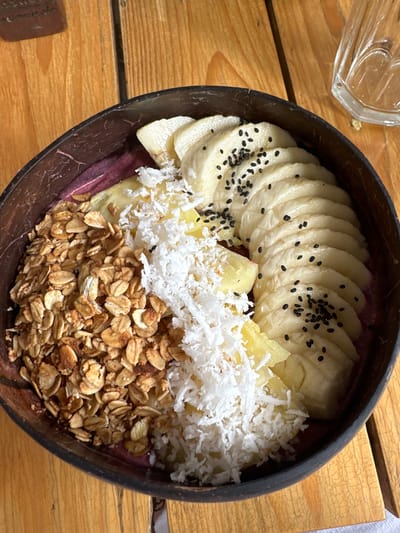 A vibrant acai bowl is served in a dark brown coconut shell. The bowl is topped with neatly arranged sliced bananas sprinkled with black seeds, chunks of pineapple, and a generous layer of shredded coconut. At the bottom, there is a portion of granola, providing a hearty, crunchy base. A glass containing an unidentifiable liquid can be seen in the corner of the image. The bowl sits on a light brown wooden table.