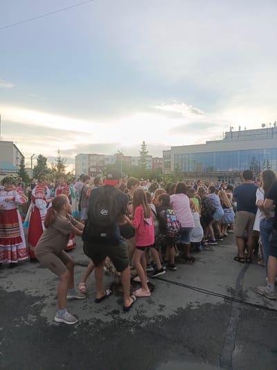 The image shows a group of people, including children and adults, participating in a tug-of-war game in an outdoor public space. The participants are gathered around a thick rope, with some individuals on both sides pulling on it. The scene appears to be a festive event, with some people wearing traditional clothing. In the background, there are multi-story buildings, suggesting an urban or town setting. The sky is partly cloudy. The ground is paved and appears wet in some areas, indicating recent rain or a water-related activity. The event is likely taking place in the city of Krivodanovka, Russia, based on the provided location information.