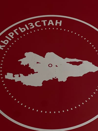 The image is a close-up of a red circular emblem with white text and a white outline of a map. The text, which appears to be in Cyrillic script, reads "КЫРГЫЗСТАН" (Kyrgyzstan) around the top arc of the circle. A dashed white line forms the inner boundary of the text. Centered within the circle is a white silhouette of the geographical shape of Kyrgyzstan. A small, dark circle is visible on the map silhouette, potentially indicating a capital or significant city. The background is a solid, deep red. There are no people, animals, or other objects visible in the image. The location is not discernible from the image itself, but the text clearly indicates it relates to Kyrgyzstan.