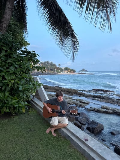 The image captures a person sitting on a concrete ledge by the ocean in Goviyapana, Sri Lanka. The subject, a young man with brown hair, is holding an acoustic guitar and appears to be playing it. He is wearing a dark t-shirt and light-colored shorts, with his bare feet visible. The setting is a scenic coastal area with rocky shores and the ocean. Waves are crashing against the rocks, and a beach with vegetation and buildings is visible in the distance. Palm fronds frame the top left of the image, suggesting a tropical environment. A small glass with a purple liquid is placed on the ledge next to the man. The sky is a clear blue with a few wispy clouds, indicating it is likely daytime, possibly late afternoon or early evening due to the soft lighting. The overall mood is relaxed and peaceful. The text "SUZUKI" is visible on the headstock of the guitar.
