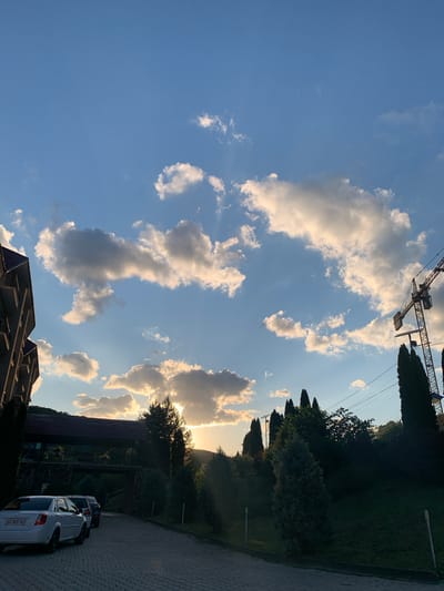 The image depicts an outdoor scene, likely in the city of Solnechnoye, Ukraine, at dusk. The sky is a vibrant blue, filled with scattered, illuminated clouds. Sunlight streams through a gap in the clouds near the horizon, casting a warm glow. In the foreground, a paved driveway is lined with parked cars. A white sedan is most prominent, with two other vehicles behind it. To the left, a portion of a multi-story building with a tiled roof is visible. Across the driveway, a bridge-like structure with a dark roof spans over a wooded area. The landscape is characterized by lush green trees and foliage, including several tall, slender cypress trees, and a construction crane is visible in the upper right portion of the image. The overall impression is one of a tranquil evening in a somewhat developed area, possibly a resort or residential complex.