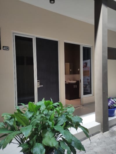 The image shows a room with a door and a window. The door is made of glass and has a white frame. It is closed and there is a screen door on the outside of it. The window is also made of glass and has a white frame. It is open. There is a plant in the foreground, and the room is in the background. The room is brightly lit and there are no people present. The number 402 is visible on the door. This room is located in Gambiran, Indonesia, at the coordinates -8.434 latitude, 114.18 longitude.