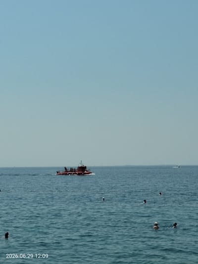 A wide shot captures a clear blue sky meeting the tranquil, rippling blue sea. A distinctive red, two-hulled vessel, possibly a tourist submarine or research craft, cruises from left to right across the mid-distance, leaving a white wake. In the foreground, several people are visible in the water, some with heads just above the surface, indicating they are swimming or wading. A rope with buoys delineates a shallow area offshore. In the far distance, another smaller boat is visible on the right horizon. The timestamp "2026.06.29 12:09" is overlaid on the lower left corner of the image. The scene suggests a typical summer day at a beach or coastal area, potentially on the island of Krk, Croatia.