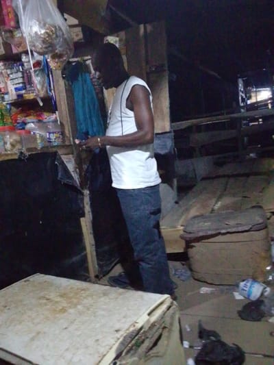 An individual wearing a white sleeveless shirt and jeans stands near a cluttered shop in Ajaokuta, Nigeria. The shop displays items such as snacks and goods hanging in plastic bags. Next to the shop, there is what appears to be a storage area with dark tarps, old furniture, and general debris on the floor. The lighting is dim.