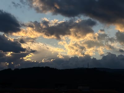 The image captures a dramatic sky with a mix of dark, heavy storm clouds and bright, sunlit clouds. A sliver of blue sky is visible between the clouds. The sun is peeking through some of the clouds, illuminating their edges with a golden glow. Below the sky is a dark silhouette of a landscape, likely a forested hill or mountain range. Two tall structures can be seen poking above the top of the hill. The foreground is very dark and contains what appears to be the tops of buildings with tiled roofs. The image was taken in Dolna Dabeva, Bulgaria.