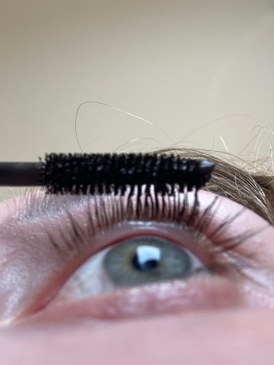 A close-up, eye-level shot shows a person's left eye as mascara is applied. The mascara wand, with its black bristles, is angled from the upper left, applying product to the upper eyelashes. The eye itself is open, revealing a light green iris with flecks of blue and a reflection of a window in the pupil. The skin around the eye shows some texture and faint redness, with a few light-colored specks that might be makeup residue. Several loose strands of light brown hair are visible near the mascara wand and above the eye, suggesting the person's hair color. The background is a plain, light-colored wall or surface, out of focus. The image does not contain any specific landmarks or visual cues that would allow for localization to Petah Tikva, Israel.