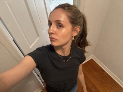A young woman with light skin and brown hair pulled into a ponytail is standing in an indoor environment, looking to her left. She is wearing a dark t-shirt and a gold necklace with two interlocking rings. She has a gold hoop earring in her left ear. The background shows a white door on the left and a wall and wooden floor on the right. The lighting appears to be artificial, creating a subtle glow on her skin. The overall scene suggests a casual, personal moment. This image does not contain enough information to determine a specific location within Miami, United States of America.
