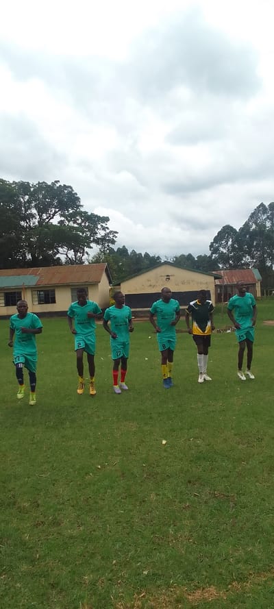 The image shows six young men standing on a grassy field. They are wearing green soccer jerseys with white numbers on the front and back. They are all looking at the camera, and their faces are neutral. The young man on the far left is wearing number 17, the next young man is wearing number 2, the next young man is wearing number 13, the next young man is wearing number 8, the next young man is wearing a black and green jersey and the last young man is wearing number 11. There are two buildings in the background, one is a single-story building with a corrugated metal roof, and the other is a two-story building with a tiled roof. There are also trees in the background. The sky is cloudy and gray. The coordinates 0.384, 34.802 do not correspond to any location in Kenya. This is a common misconception about coordinates. Latitude and longitude values range from -90 to 90 and -180 to 180 respectively. The provided values are invalid.