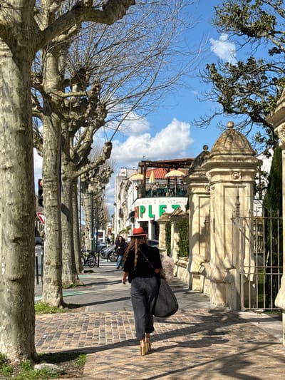 The image depicts a sunny urban street scene at 12 Place de Tourny, Bordeaux, France.

In the foreground, a person is observed from behind, walking from right to left on a cobblestone sidewalk. This individual wears a reddish-orange fedora-style hat, a dark short-sleeved top, dark blue or grey pants, and metallic gold platform high-heeled shoes. Long, wavy brown hair extends down their back. They carry a large, dark, possibly woven bag on their left shoulder.

To the left, a row of mature, pruned plane trees with bare branches lines the sidewalk, casting shadows. Further down the street, a traffic light displays a red signal, and vehicles, including a parked car and a bicycle, are visible. Other pedestrians can be seen walking in the distance.

On the right, ornate light-colored stone pillars with domed finials and an iron gate mark the entrance to a property. Beyond these, a multi-story building features a restaurant named "SIMPLEZIA," identified by a green sign with white lettering. Above it, "LA PIZZA E LA VITA" is also visible. An outdoor terrace on the building's first floor is furnished with tables, chairs, and several yellow sun umbrellas. The sky is blue with scattered white clouds, and bright sunlight creates distinct shadows across the scene.