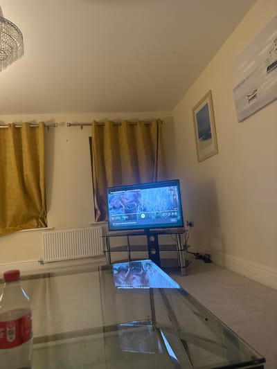 The image shows an interior scene. In the foreground is a glass coffee table. A bottle of water is partially visible on the table. A television is positioned on a stand against a wall, displaying what appears to be a video. Behind the TV, curtains hang on either side of a wall-mounted radiator. Two framed pictures hang high on the wall. The image is rotated counter-clockwise.
