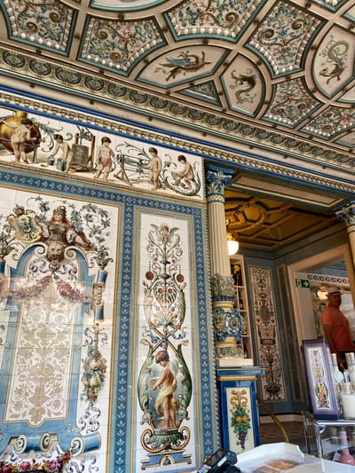 The image shows the ceiling and walls of Pfunds Molkerei in Dresden, Germany. The walls and ceiling are covered in colorful, decorative tiles with various patterns, including floral motifs, mythical creatures, and depictions of people. The color palette consists of blues, greens, and creams. Some surfaces are reflective. The angle of the photograph is upwards, capturing the ornate details. Part of a column is visible. In the upper part of the image, a reflection shows a person wearing a peach-colored shirt, merchandise, and an exit sign with a green running figure.
