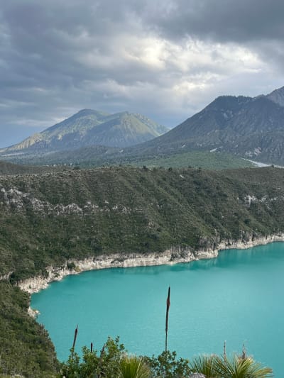 The image captures a panoramic view of a vibrant turquoise lake, likely within or near Guadalupe Victoria, Mexico. The lake's edge is bordered by steep, rocky cliffs covered in dense green vegetation, featuring a distinct white band running horizontally along the cliff face. Beyond the lake and cliffs, a series of layered mountains rise, their slopes varying in color from dark green to lighter, barren tones, all under a dramatic sky filled with heavy, gray clouds tinged with sunlight breaking through. In the foreground, the lake's surface is smooth and reflective, with a few tall, thin plants emerging from the water near the shore. Lush green foliage, including palm-like fronds, occupies the lower edge of the frame, suggesting a natural and somewhat wild environment. There are no people or animals visible in the scene.