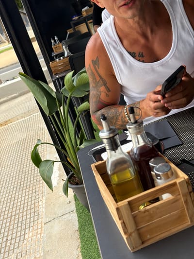 The image shows a man wearing a white tank top and holding a smartphone in his left hand. He has tattoos on his arms and chest. In front of him, on a dark table, is a wooden crate containing two glass bottles with dispensers, one filled with a yellow liquid (likely olive oil) and the other with a dark liquid (likely balsamic vinegar). There are also two silver salt and pepper shakers. A black napkin is placed to the right of the crate. A potted plant with large green leaves is visible to the left of the table. The background shows a street or sidewalk with a diamond-patterned paving and a black railing, suggesting an outdoor setting. The man is at a table, likely in a restaurant or cafe, in the city of La Vila Joiosa, Spain.