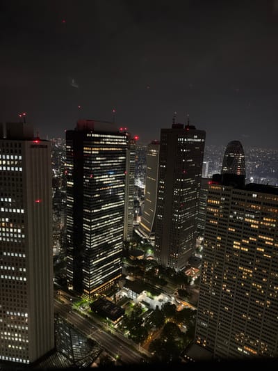 This is an aerial, nighttime photograph of the Shinjuku district in Tokyo, Japan. The image showcases a cluster of illuminated skyscrapers, with their windows emitting a warm, yellow glow against the dark sky. Several tall buildings dominate the foreground and midground. To the left is a lighter-colored, rectangular skyscraper with a grid of lit windows. Adjacent to it, to the right, is a darker, taller skyscraper also filled with glowing windows. Further to the right, another large, dark skyscraper is visible, and a distinctive building with a curved, lattice-like facade stands out in the background. The cityscape stretches out into the distance, with the lights of the city creating a hazy glow. Roads and smaller buildings can be seen at the base of the skyscrapers, with streetlights and the lights from these structures illuminating the ground level. Trees are also visible, adding a touch of nature to the urban landscape. Red aviation lights are present on the tops of some of the buildings. The overall impression is of a bustling, modern metropolis at night.