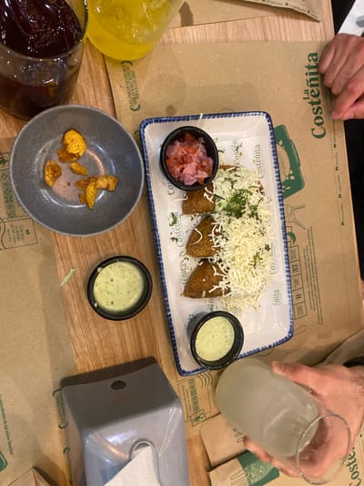 The image shows a table setting in a restaurant, presumably in La Aurora, Ecuador given the references to "La Costeñita" on the placemats and plates. Two people are visible; one is holding a glass of a clear beverage with ice, while the hands of another person are near the top of the frame.
The table is covered with paper placemats bearing the La Costeñita logo and branding. At the center of the table, a rectangular plate features food items, including what appears to be arepas topped with shredded cheese and herbs. Beside this, are two small bowls containing salsa or a similar condiment. A gray bowl contains what seem to be fried plantain chips. Two smaller bowls, also in gray, hold light green dipping sauce.
A glass of a dark beverage and a yellow drink are visible on the left, along with a gray tissue box on the right side of the table. The scene suggests a casual dining experience.