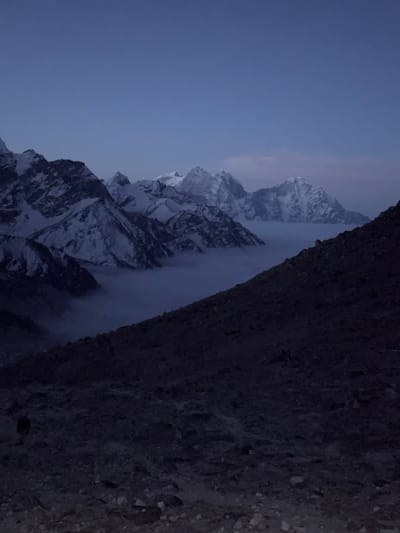 The image captures a dramatic, high-altitude mountainous landscape, likely at dawn or dusk, in Khumjung, Nepal. The photo is oriented vertically, showing a dark, textured slope or rock face in the foreground on the right side. This foreground element is composed of rough, uneven ground or rock, appearing dark and silhouetted against the distant view.
Beyond the immediate foreground, a majestic range of snow-capped mountains dominates the scene. Multiple jagged peaks, covered in white snow and ice, stretch across the middle ground and background, their forms partially obscured by atmospheric mist or low-hanging clouds that fill the valleys and lower slopes. The dim, diffused light suggests either very early morning before sunrise or late evening after sunset, creating a serene and somewhat somber mood. The sky above is a deep, purplish-blue, indicating twilight conditions, with faint, star-like specks visible in the upper reaches.
The scene conveys the immense scale and rugged beauty of the Himalayan terrain. There are no identifiable people or man-made objects. The weather appears cold and clear above the clouds, with mist clinging to the mountain bases. No visible text is present.