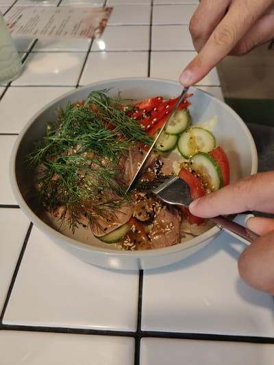 A close-up shot shows hands using a fork and knife to cut into a salad in a light grey bowl. The salad contains slices of meat, dill, sliced cucumbers, sliced red bell peppers, and sesame seeds, all coated in a glistening dressing. The bowl sits on a white tiled surface. In the upper left background, a menu or pamphlet is visible. The overall scene is well-lit, suggesting an indoor dining setting. There is no information in the image to suggest the location is in Кварса, Russia.