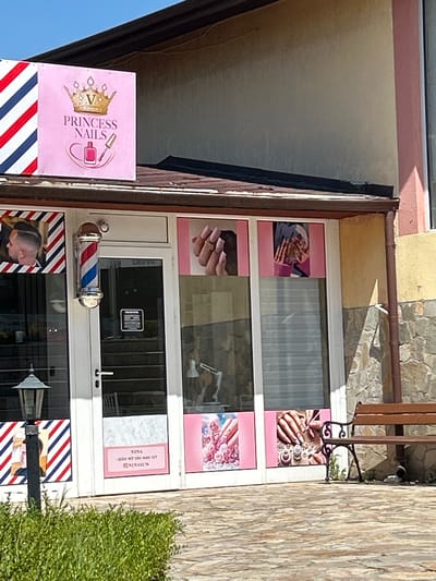 An exterior shot captures a commercial establishment with a prominent pink sign featuring a golden crown and the text "PRINCESS NAILS" with an image of a nail polish bottle and brush. The building appears to be a nail salon, evidenced by several window displays showcasing manicured hands and nails. To the left, a barber pole and a framed image of a man's haircut suggest a neighboring barbershop. A glass-paned door with a handle and a sign are visible. The facade is a mix of yellow stucco and stone. A wooden bench sits outside the establishment, and lush green bushes are present in the foreground. The sky is clear and blue, indicating a sunny day. The location is identified as Aksakovo, Bulgaria.