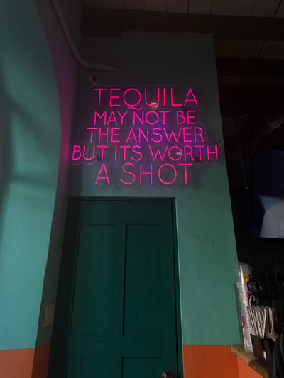 The image presents an interior scene within what appears to be a bar or restaurant in San José del Cabo, Mexico. A prominent, illuminated pink neon sign is mounted on a two-toned wall, displaying the text: "TEQUILA MAY NOT BE THE ANSWER BUT ITS WORTH A SHOT." The wall's upper section is painted a light teal-green, while the lower portion is orange. Centered beneath the neon sign is a dark teal-green wooden door, featuring two vertical panels and equipped with a round metallic doorknob. To the right of the door and extending into the mid-ground, a bar service area is visible. This area includes a counter holding stacked clear plastic cups and a container with straws. Behind these items, dark bottles, likely spirits, are arranged on shelves, and a section of a television screen displaying a blurred image of a person's face is present. A dark-colored fan is partially visible in the upper right corner. No individuals are explicitly depicted in the scene; the ambient lighting is dim, with the neon sign serving as a primary light source.
