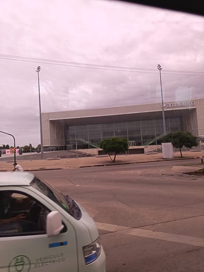 The image shows Antel Arena, a large indoor arena in Montevideo, Uruguay. The arena has a glass facade and the words "antel arena" are visible on the top right. Two tall light posts stand in front of the building. In the foreground, a light-colored electric vehicle, possibly a delivery van, is visible on the street. A person is inside the vehicle. The sky is overcast. Other trees and street infrastructure are visible in the background.