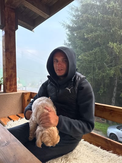 A man wearing a black hooded jacket and a black vest sits on a wooden bench with a fluffy light brown dog in his lap. He is outdoors on a covered porch, with wooden beams above and a wooden railing. The background shows trees covered in mist, a few buildings with green roofs, power lines, and a silver car. The environment appears to be the village of Trešnjevik, Montenegro, characterized by its mountainous terrain and often misty weather conditions. The man's gaze is directed towards the camera, and he appears to be holding the dog gently. A white fluffy rug is visible on the bench beneath the man and dog.